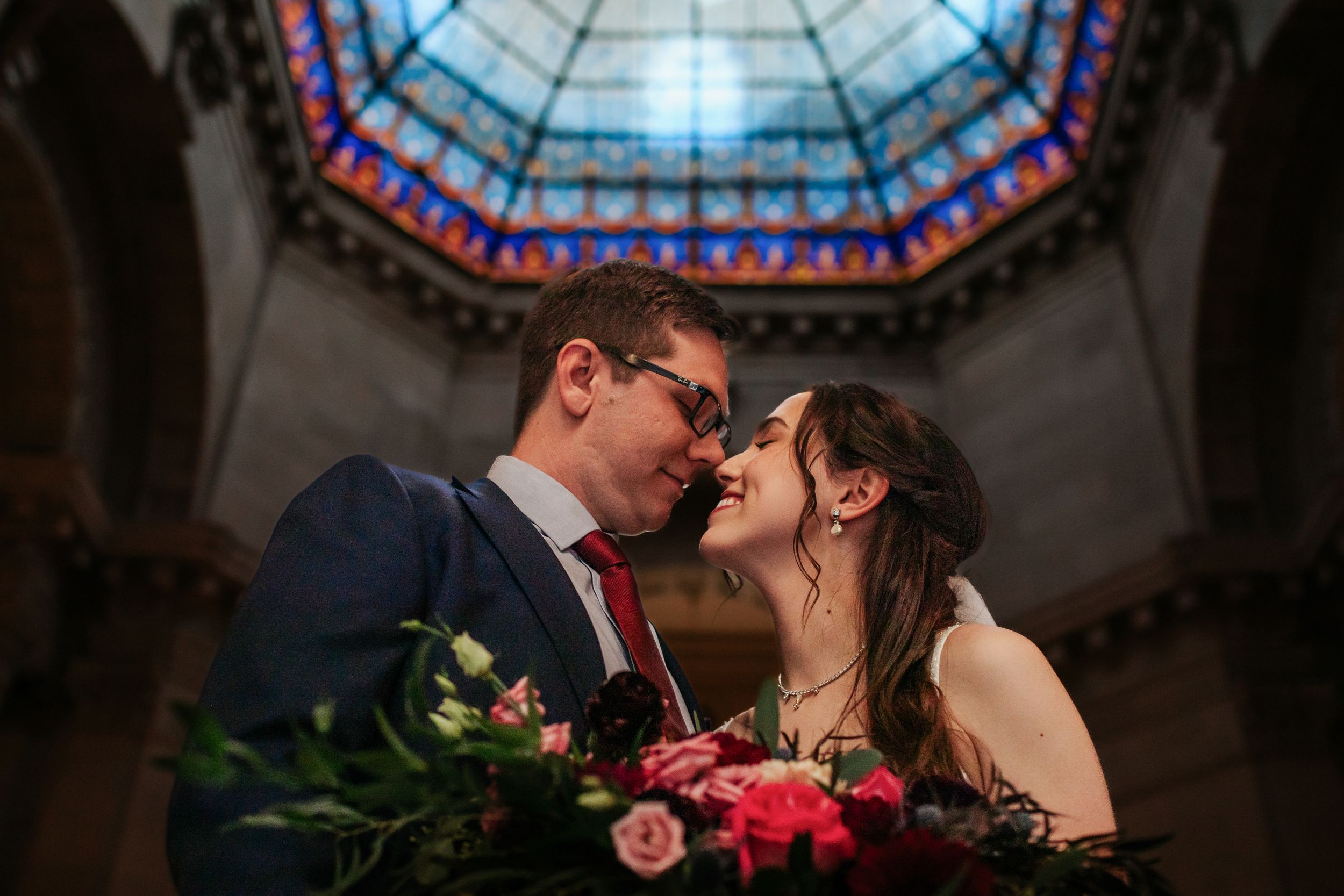 Wedding couple poses beneath stunning stained glass ceiling in historic building interior.