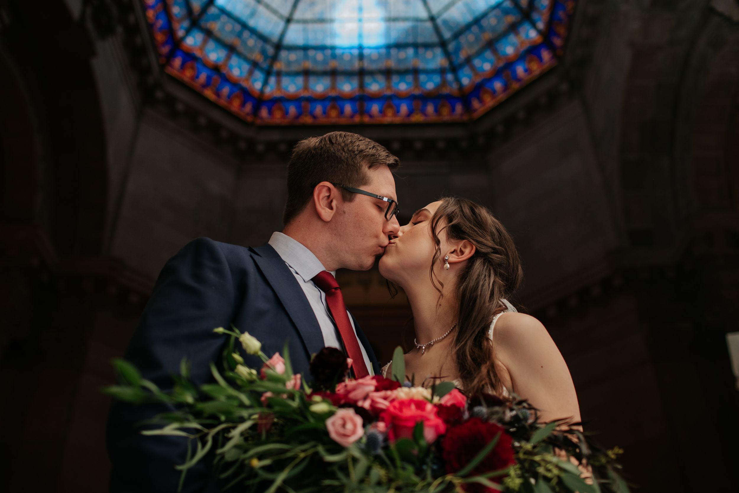 Wedding couple embraces under a decorative stained glass dome in an elegant historic building.