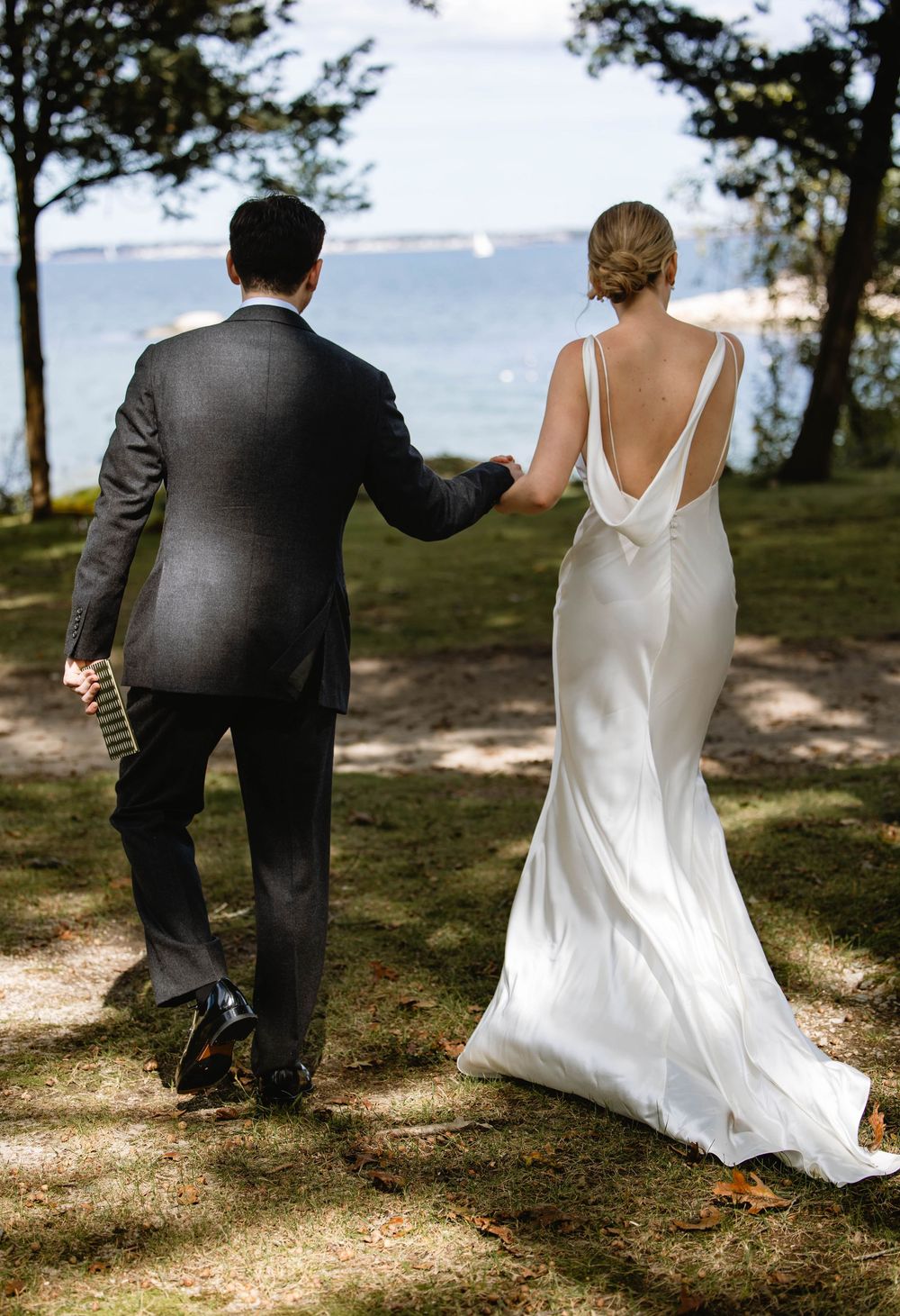 A couple in formal attire walks along a waterfront path with sunlight filtering through trees. Weatherlow Farm wedding. Westport Mass