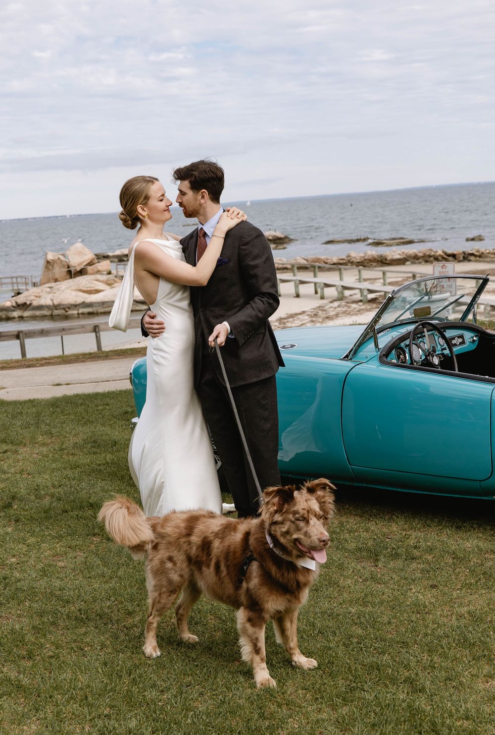 A couple poses with their dog next to a vintage turquoise car by the ocean. Weatherlow Farm wedding. Westport Massachusetts. New England.
