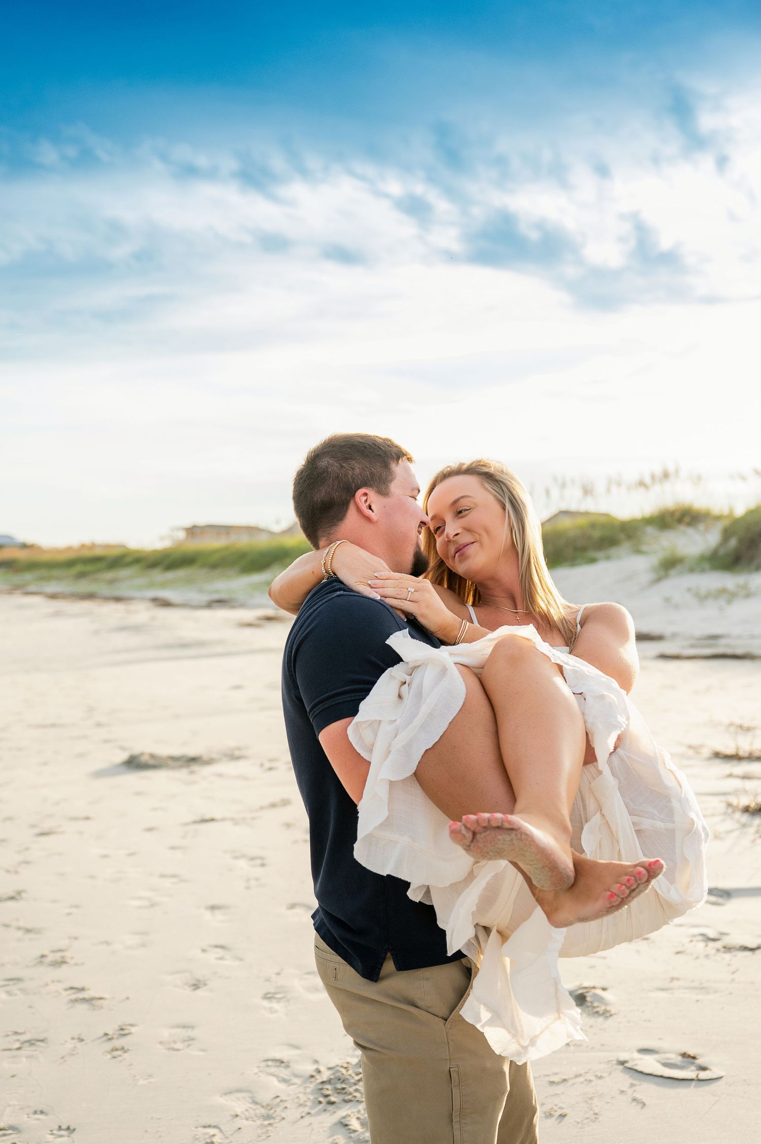 Playful beach engagement photos with couple in white and khaki against sand dunes and blue sky.