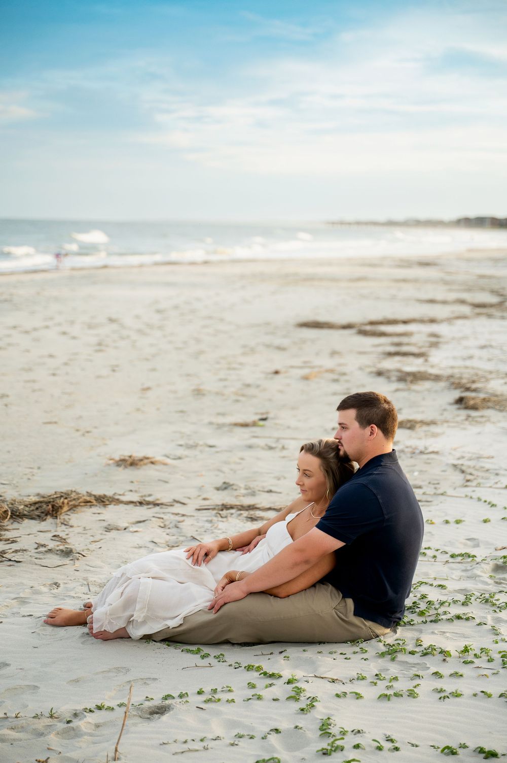 Couple relaxing on beach sand with scattered seaweed during romantic photoshoot.