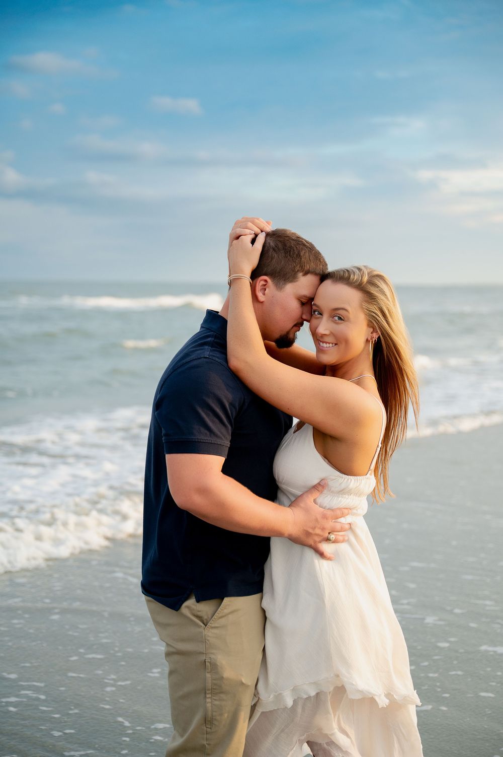 A romantic couple embraces on a beach at sunset with ocean waves in the background.