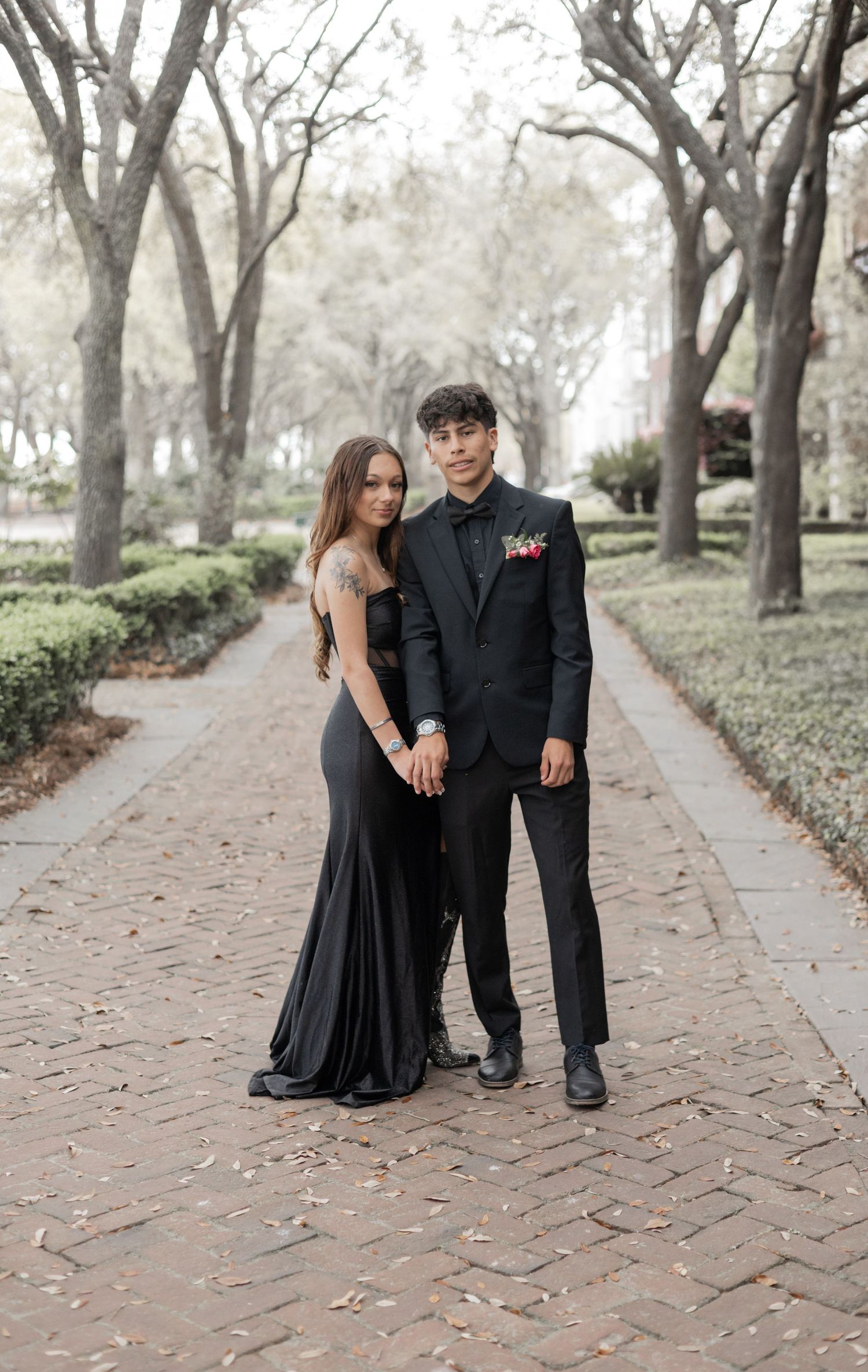 A couple in formal attire poses together on a brick pathway surrounded by bare winter trees.