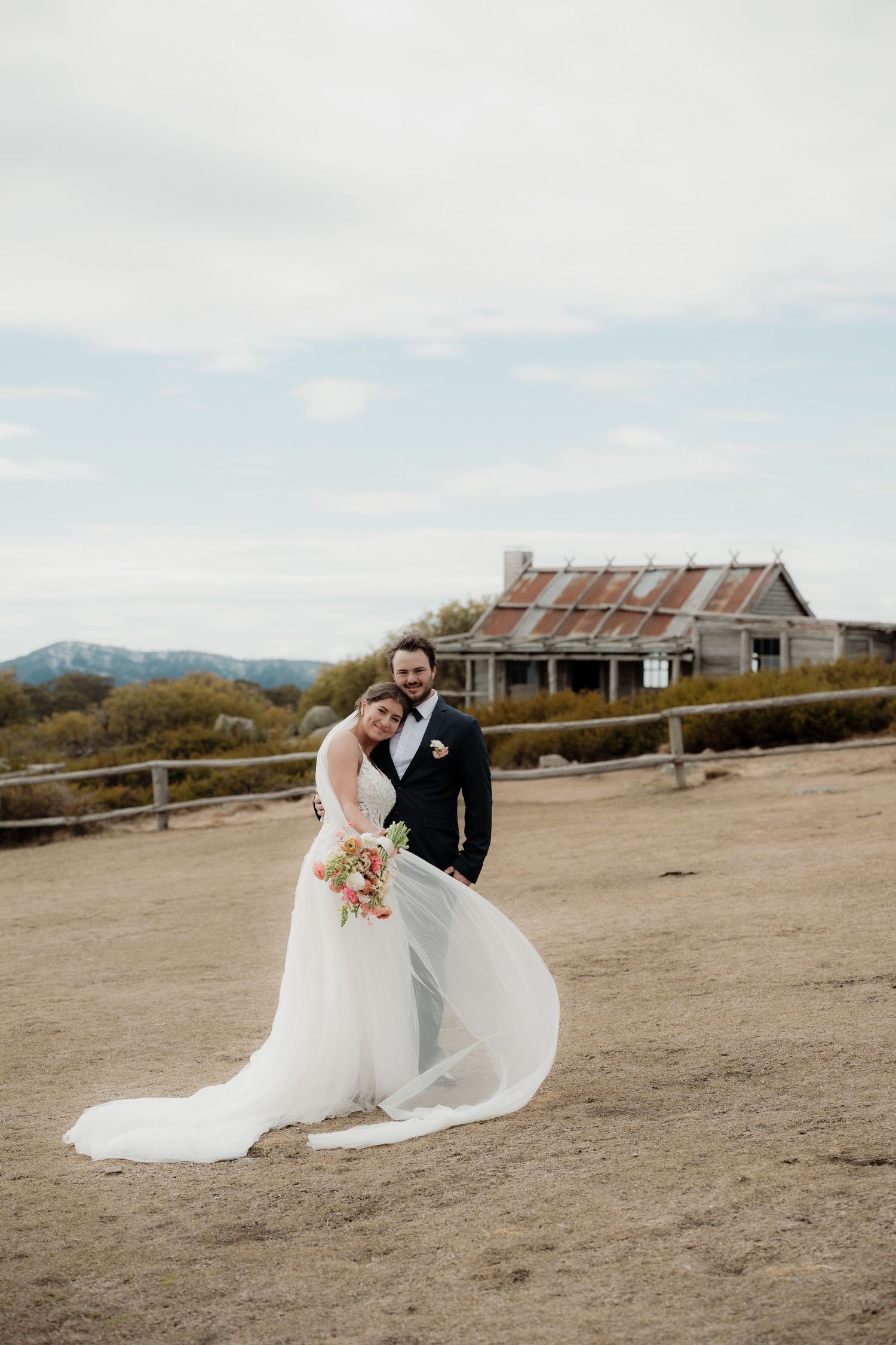 A couple in wedding attire shares an intimate moment on a scenic hilltop with rustic buildings in the background at Craigs Hut