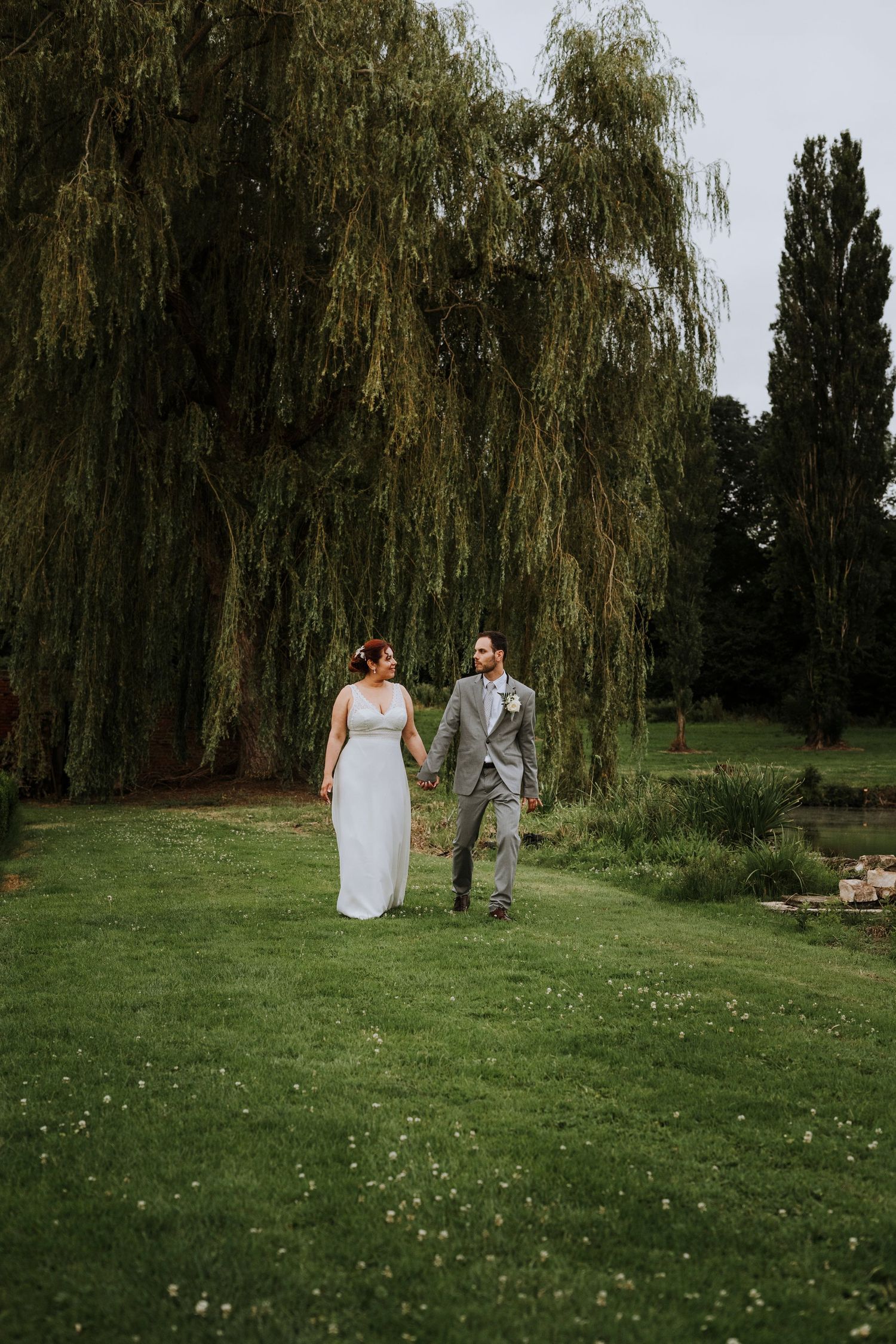 A couple in wedding attire walks together on a lush green lawn with weeping willow trees in the background.