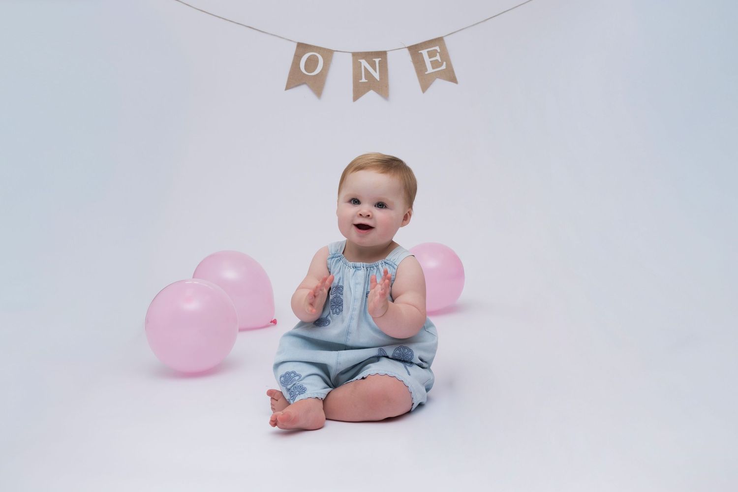 Baby girl celebrating first birthday surrounded by pink balloons during cake smash photo session