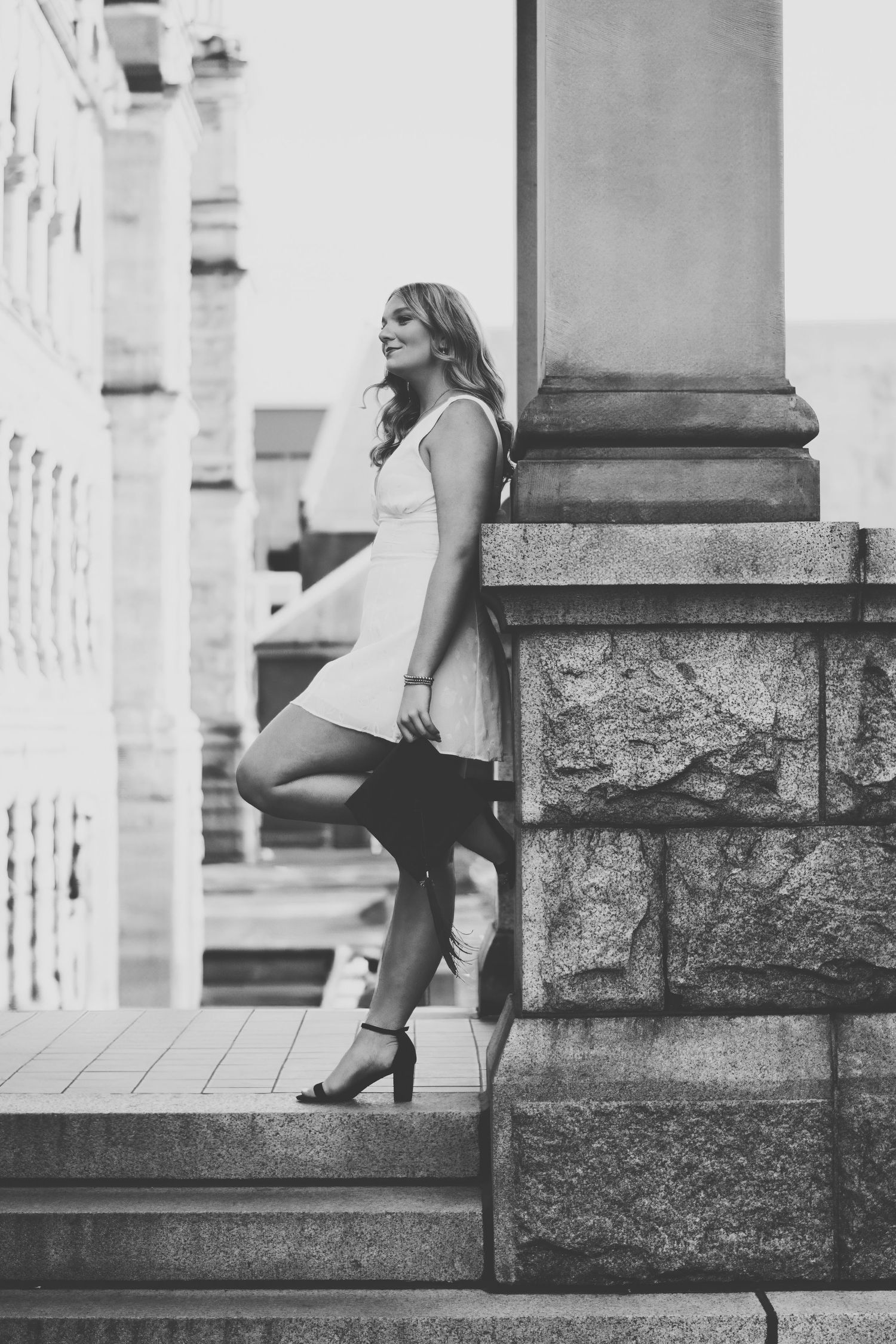 Black and white photo of someone in a short white dress posing on stone steps near classical architecture columns.