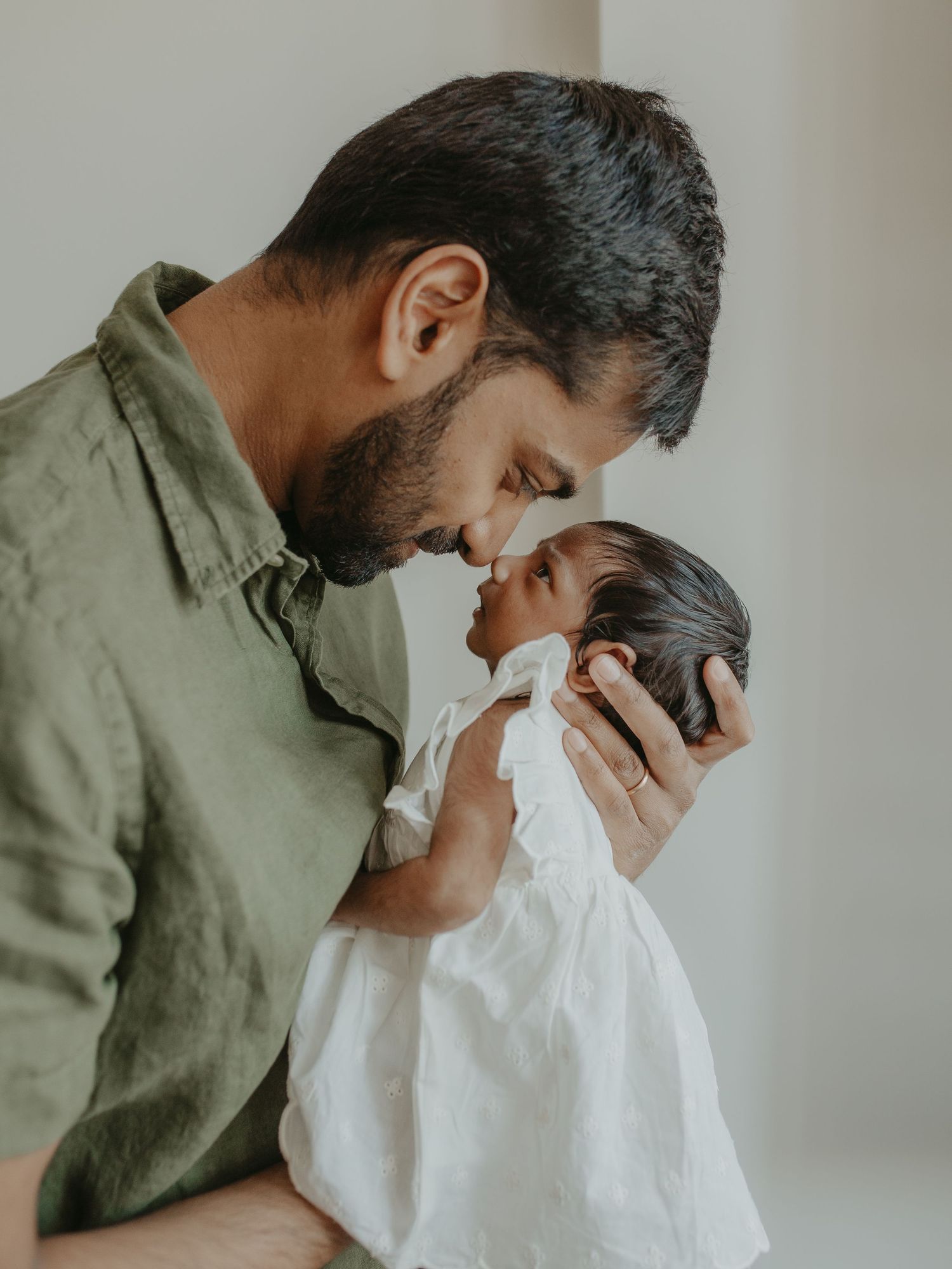 A bearded father in a green shirt tenderly touches noses with his newborn baby dressed in white.