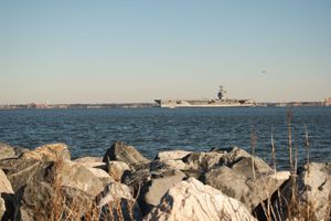 Large rocks line a coastal shoreline with calm blue water and distant buildings visible on the horizon.
