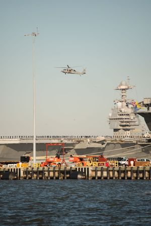 Military helicopters hover above a cruise ship docked at a waterfront pier with orange rescue boats nearby.