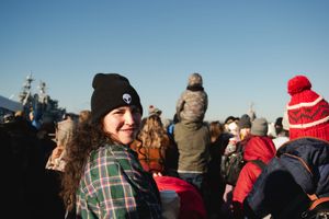 A crowd of spectators gathers outdoors wearing winter hats and coats while watching an event on a sunny day.