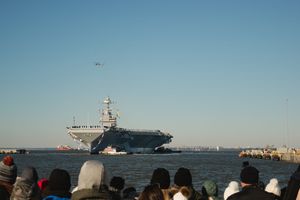 Crowd watches as large aircraft carrier ship navigates through harbor waters under clear blue sky.