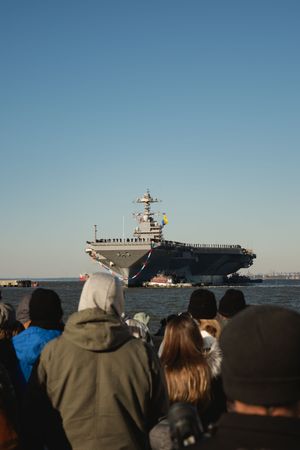 A crowd of spectators watches an aircraft carrier on the horizon against a clear blue sky.