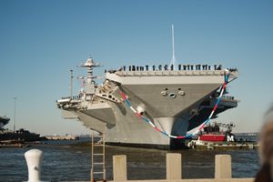 Military aircraft carrier docked at port with support vessels and equipment visible along its deck.