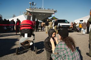 People gather outdoors at a public event with vehicles and tents visible against a bright blue sky.