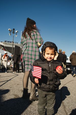A young child in winter clothing holds a small American flag at an outdoor gathering on a sunny day.