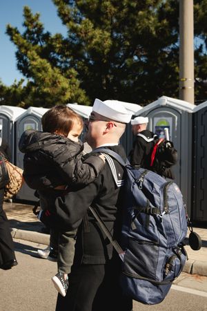 People line up near portable toilets and flagpoles in an outdoor area.