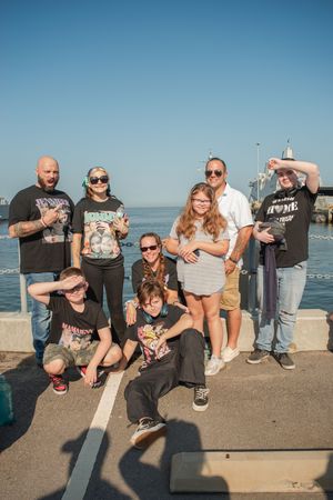 A group poses together on a concrete pier by the ocean on a sunny day, wearing casual attire and sunglasses.