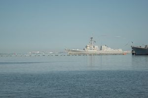 A military naval vessel sails across calm blue waters with a distant shoreline visible on the horizon.