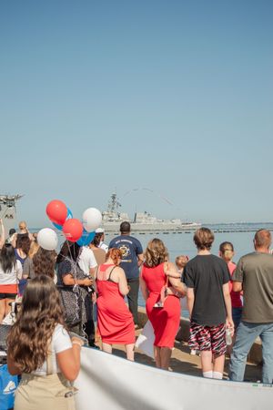 A crowd gathers at a waterfront event holding red and white balloons while looking out at the harbor.