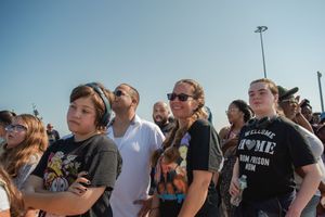 Concert crowd stands outdoors watching performance during daytime festival.
