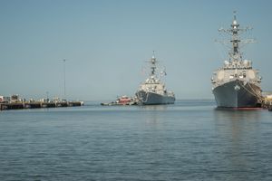 Two naval warships docked in a calm harbor on a clear day with blue skies.