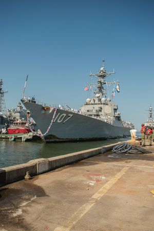 Navy warship docked at port with clear blue sky and multiple vessels visible in the background.