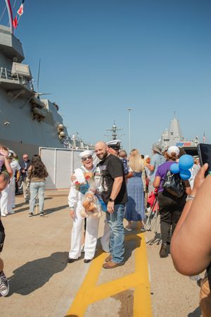 A crowd of visitors gathers on a naval ship deck with yellow safety lines under clear blue skies.