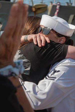 A sailor in a white uniform embraces someone in a tender moment during what appears to be a homecoming.