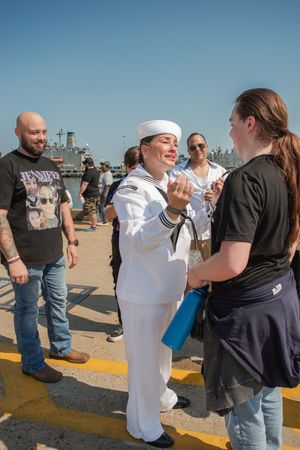 Sailors and civilians interacting on the deck of a military ship during a sunny day celebration.