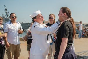 A group of people interacting on a sunny dock with naval vessels in the background.