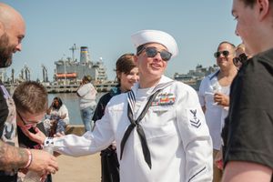 A group of sailors in white uniforms gather outdoors near a waterfront on a sunny day.