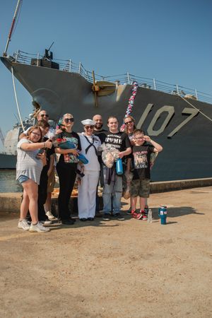 A group poses together in front of a large naval ship numbered 107 on a sunny day.