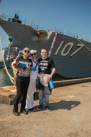Several people gathered for a photo beside a large military vessel marked 107 in bright daylight.