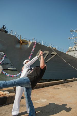 Two people dance in front of a large naval ship decorated with patriotic bunting at a dock.