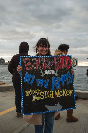 Someone holds a handmade welcome home sign with blue and red lettering on a pier near the water at sunset.