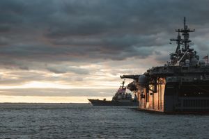 A naval warship silhouetted against a dramatic cloudy sunset sky on the ocean.