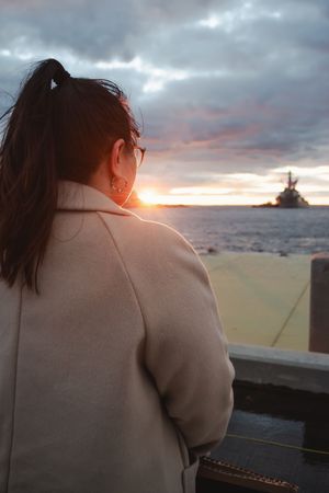 A figure in a beige coat looks out over a scenic landscape at sunset, creating a contemplative silhouette against the sky.