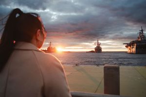 Someone watches a dramatic sunset over the ocean with silhouetted naval ships on the horizon.