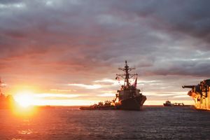 Navy warship silhouetted against a dramatic orange sunset sky over calm ocean waters.