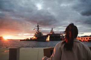 Someone watches a dramatic sunset over naval warships docked at port with stormy clouds overhead.
