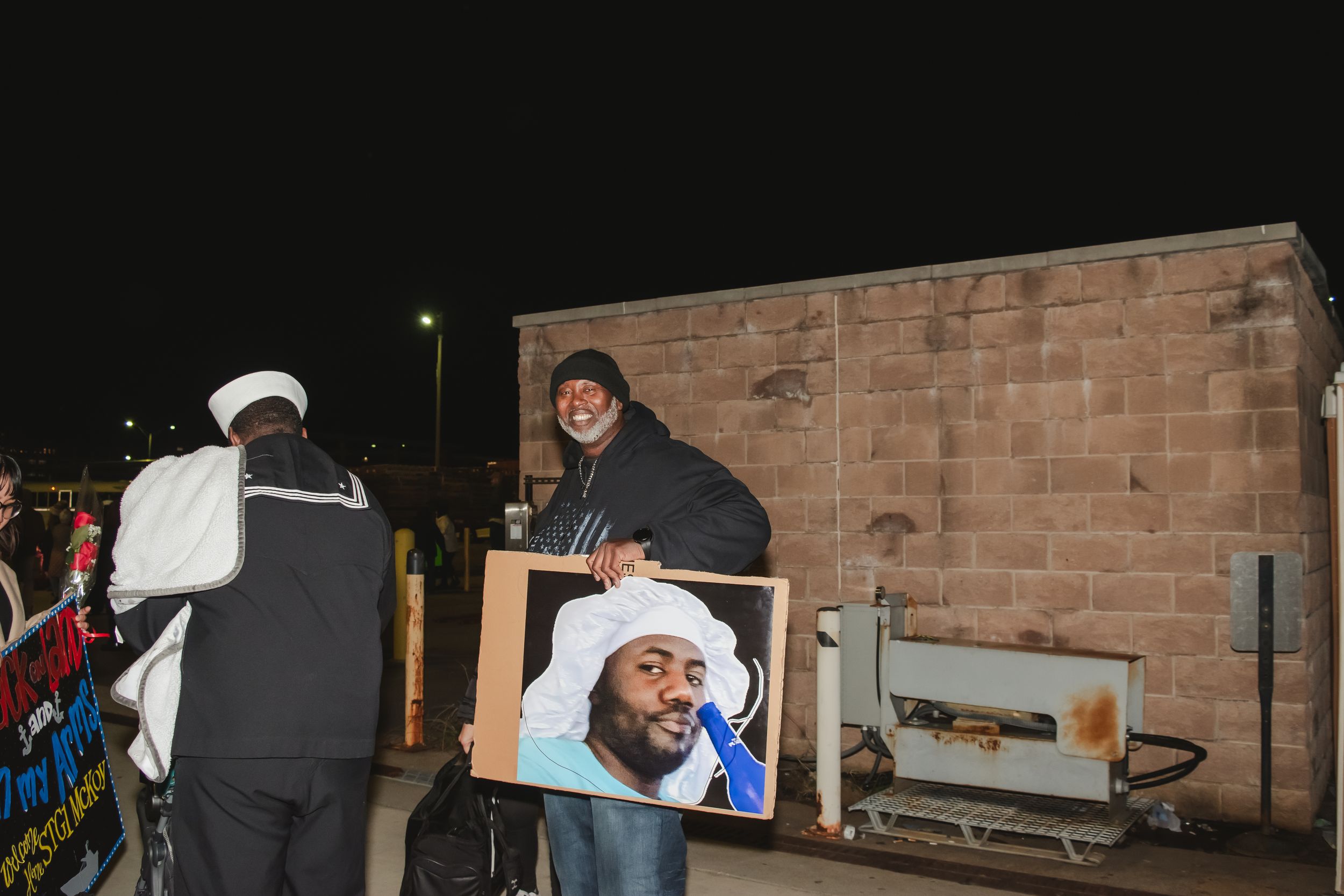 People gathered outside at night near a brick wall with a framed photo display.