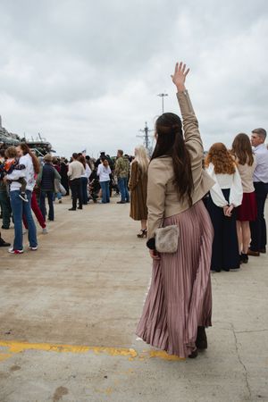 A crowd gathers on a pier with someone in a pleated mauve skirt and beige jacket waving to a naval ship.