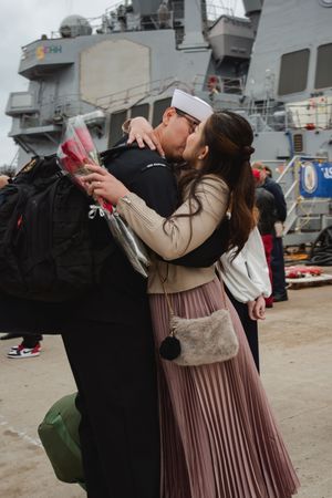 A sailor and loved one share an emotional embrace in front of a naval ship during a homecoming ceremony.