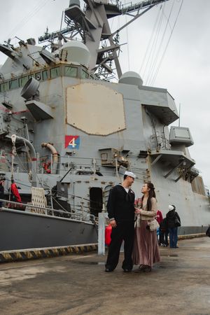 A couple shares a romantic moment in front of a naval ship docked at port during a special occasion.