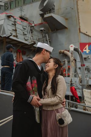 A tender moment between a couple beside a large military vessel at a naval dock.