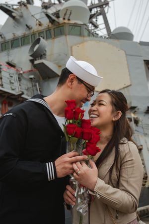 A romantic moment unfolds on the dock with red roses against a naval ship backdrop.