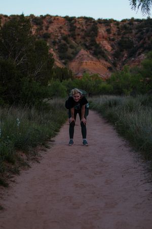 A person bends down on a dirt hiking trail surrounded by red rock cliffs and desert vegetation at dusk.