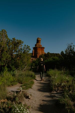 A hiker walks along a sandy trail towards red rock formations in a desert landscape under blue skies.